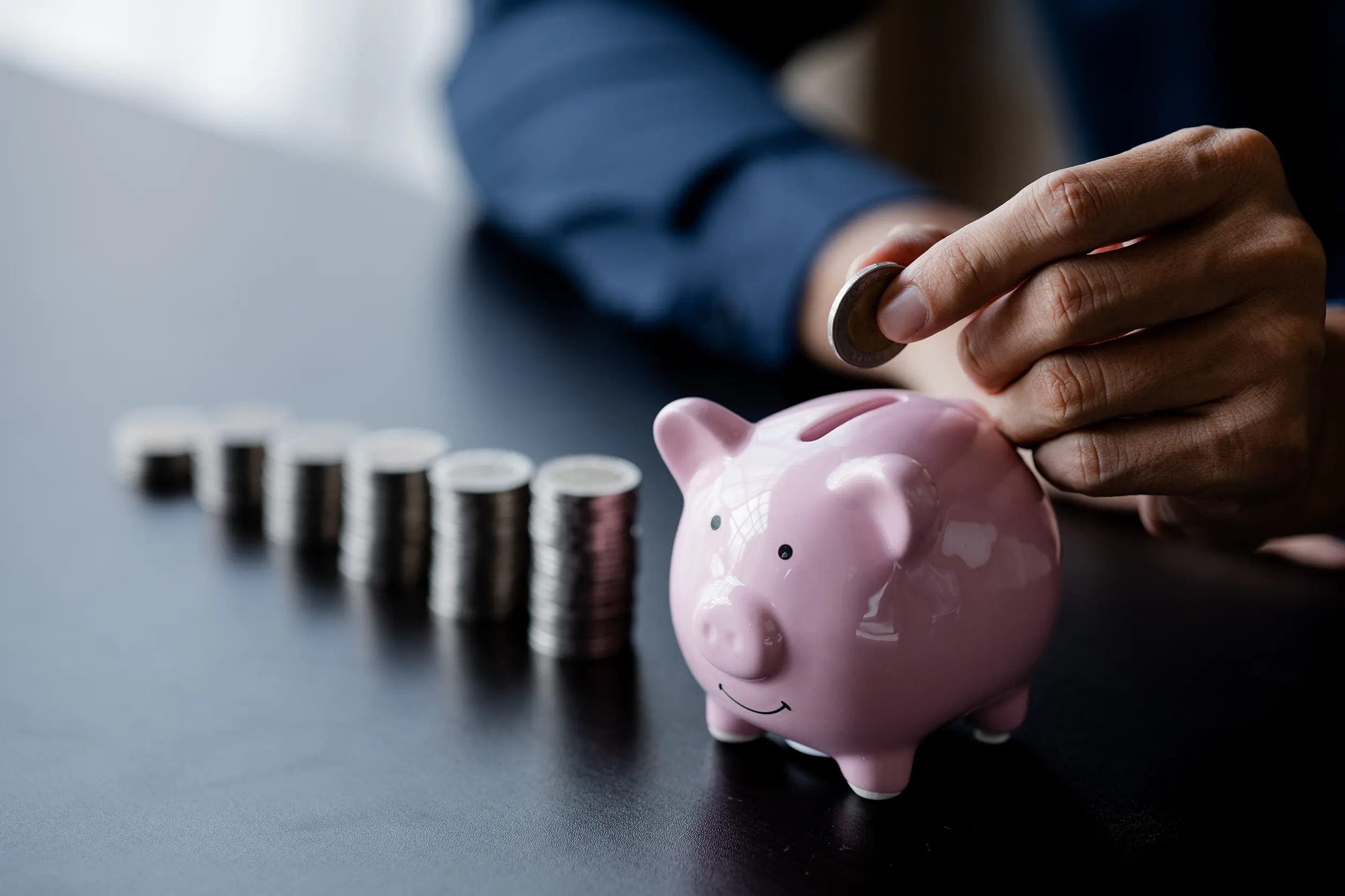 stacks of coins leading to a small pink piggy bank with man inserting coin